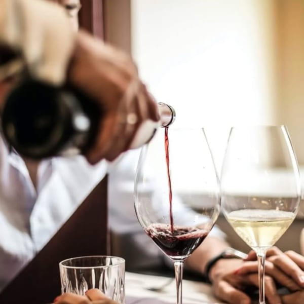 Red wine being poured into a glass, with another glass of white wine and hands visible.
