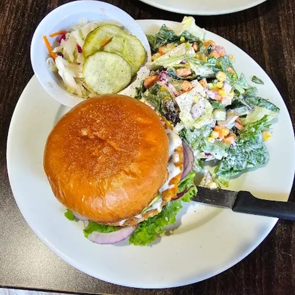 A chicken sandwich with lettuce, onion, and sauce, served with a side salad and pickles.