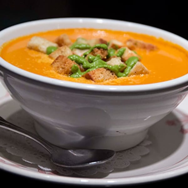 A white bowl of orange tomato soup topped with croutons and green pesto, with a spoon.
