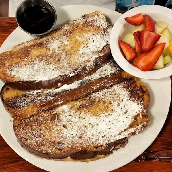 Two slices of French toast with powdered sugar, a side of fruit, and syrup on a white plate.