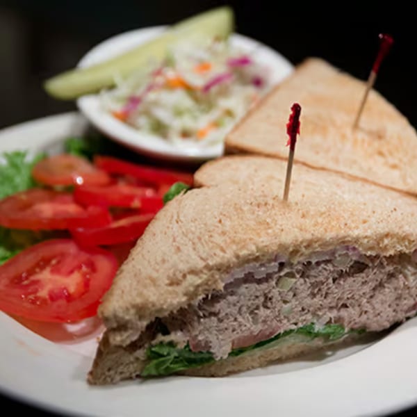 A tuna sandwich with lettuce and tomato, served with coleslaw and a pickle spear.