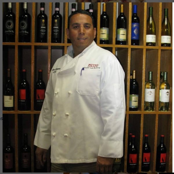 Misto Caffe & Bakery chef in a white uniform stands in front of a wine rack.