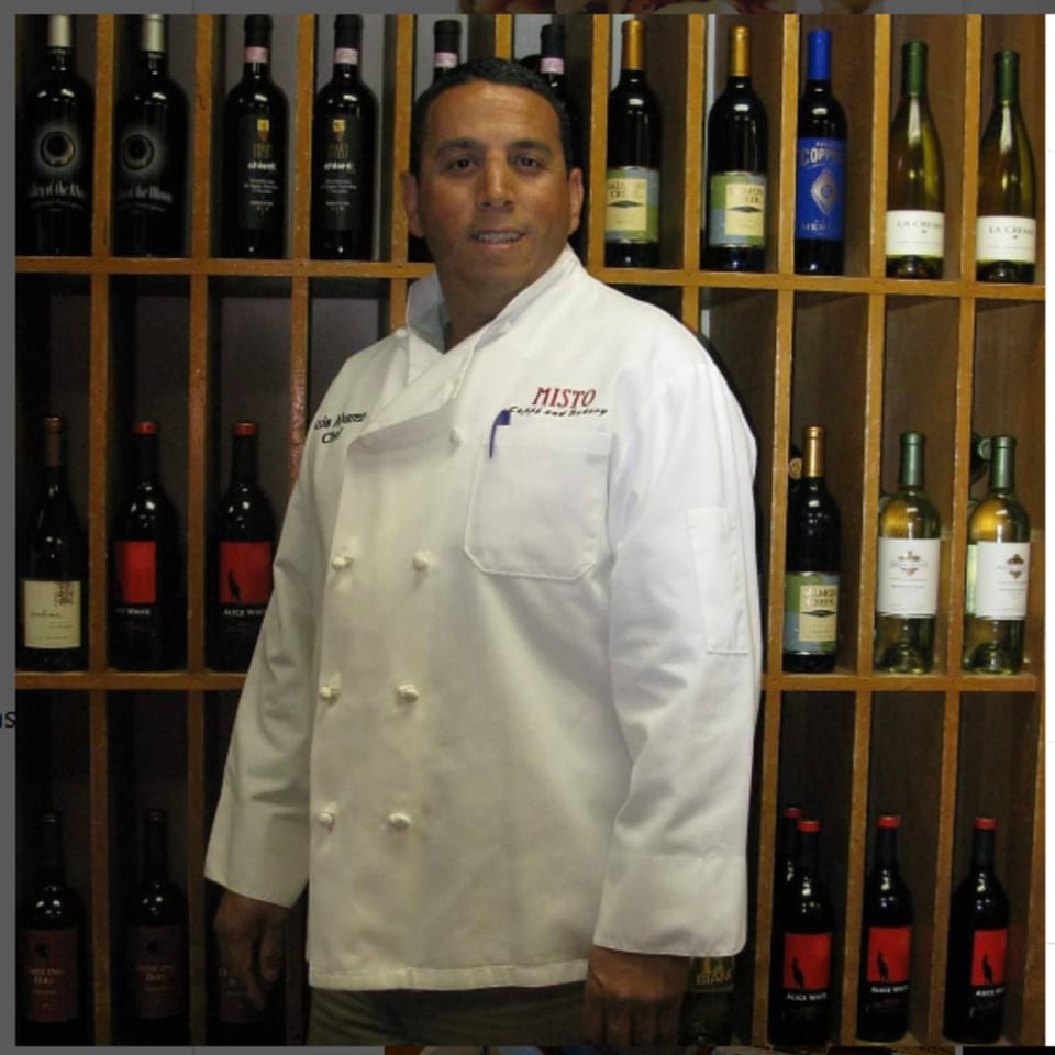 Misto Caffe & Bakery chef in a white uniform stands in front of a wine rack.